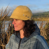 A woman wearing a mustard yellow wool cap with metal snap buttons, standing outdoors near a body of water with tall reeds in the background.