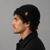 A man in profile wearing a black knit beanie with a small tan leather brand patch on the side, photographed against a plain gray studio background.