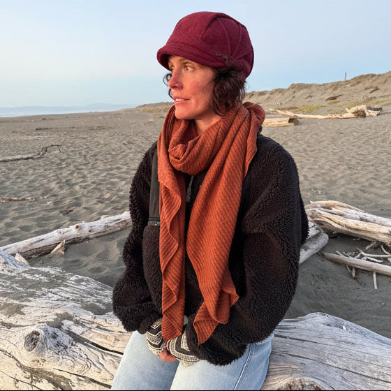 A woman sitting on a piece of driftwood on a sandy beach, wearing a dark brown sherpa fleece jacket, a rust-orange waffle-knit scarf, a burgundy knit cap, and light-wash blue jeans.