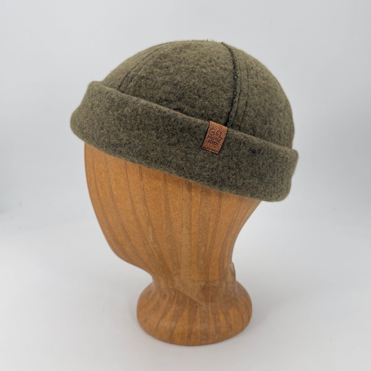 An olive green felt knit beanie hat displayed on a light brown wooden mannequin head against a plain white studio background.