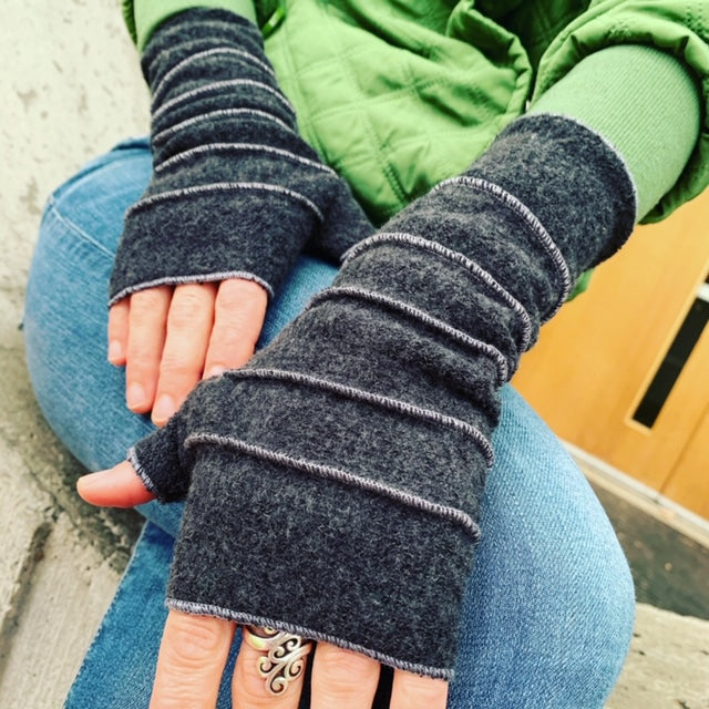 A close-up of a person wearing dark charcoal gray fingerless knit gloves with horizontal ribbed stitching, paired with blue jeans and a green jacket.