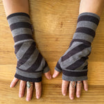 A pair of fingerless knit arm warmers with horizontal dark brown and gray stripes, worn on a person's hands over a wooden tabletop.