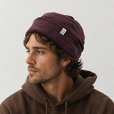 A man wearing a dark plum-colored slouchy beanie with a small white tag and a brown hoodie, shown in profile against a light gray background.