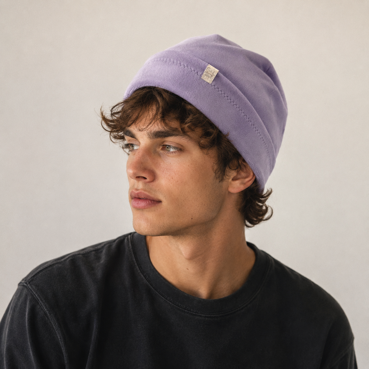 A man wearing a light purple knit beanie and a black crewneck t-shirt, photographed against a neutral light gray background.