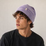 A man wearing a light purple knit beanie and a black crewneck t-shirt, photographed against a neutral light gray background.