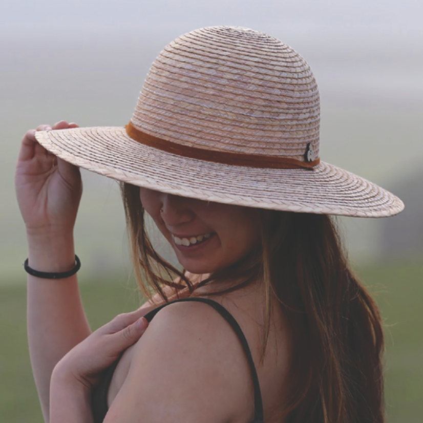 Female model blurred background wearing Anya Palm Straw Sun Hat, featuring wide brim sun protection and adjustable strap.