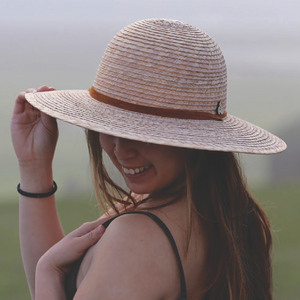 Female model blurred background wearing Anya Palm Straw Sun Hat, featuring wide brim sun protection and adjustable strap.