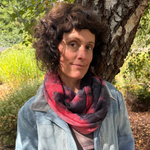 A woman with curly brown hair stands outdoors in front of a tree, wearing a light blue denim jacket, a red and black patterned scarf, and beige pants.