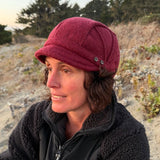 A woman wearing a dark red newsboy cap and a black fleece jacket, sitting outdoors on a sandy dune with trees in the background.