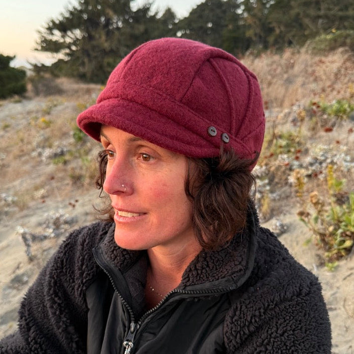 A woman wearing a dark red newsboy cap and a black fleece jacket, sitting outdoors on a sandy dune with trees in the background.