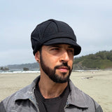 A man standing on a sandy beach wearing a black cap, a dark gray overshirt over a black t-shirt, with ocean waves, rocks, and a forested hill in the background.
