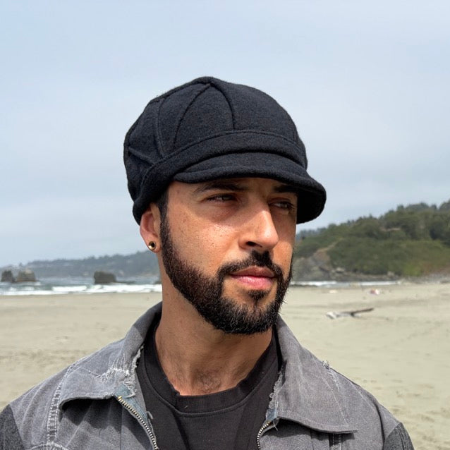 A man standing on a sandy beach wearing a black cap, a dark gray overshirt over a black t-shirt, with ocean waves, rocks, and a forested hill in the background.