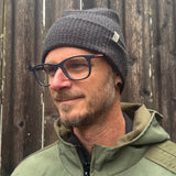 Man wearing a gray knit beanie and green jacket against a wooden fence.