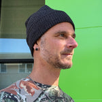 Man wearing a black knit beanie and camouflage shirt against a green wall.